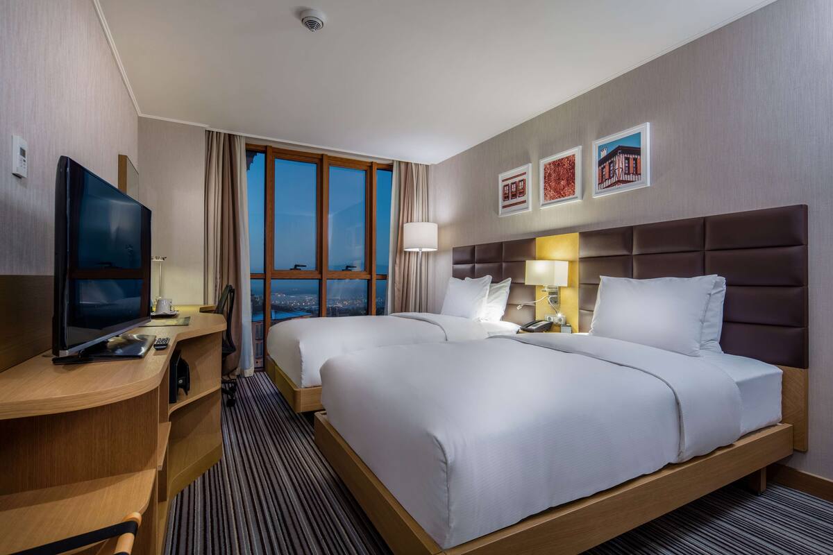 A hotel room with two beds, a desk with a TV, a lamp, and a view of the cityscape.