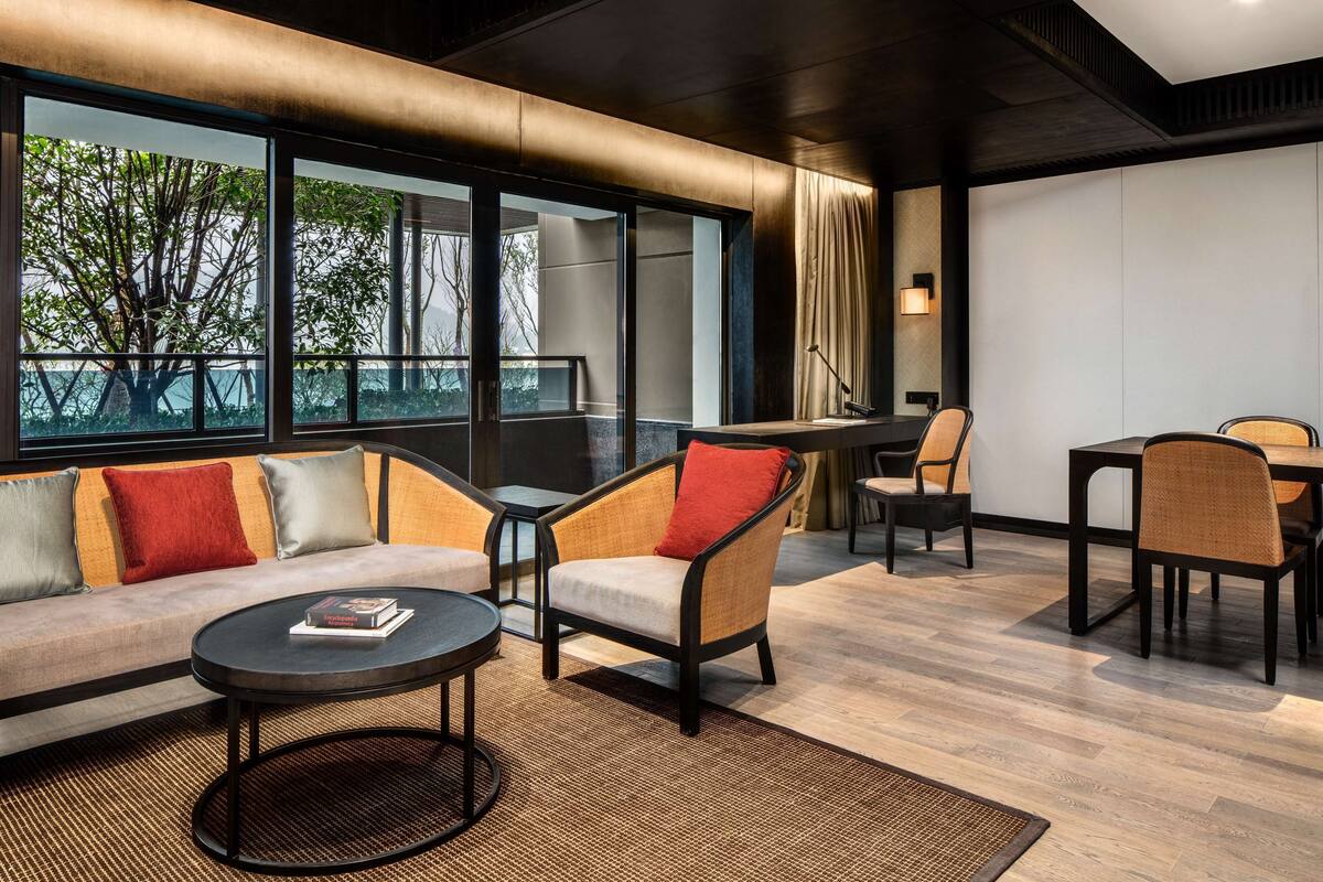 Habitación de hotel moderna con sofá, sillones, una mesa de comedor y un amplio ventanal con vistas a árboles.