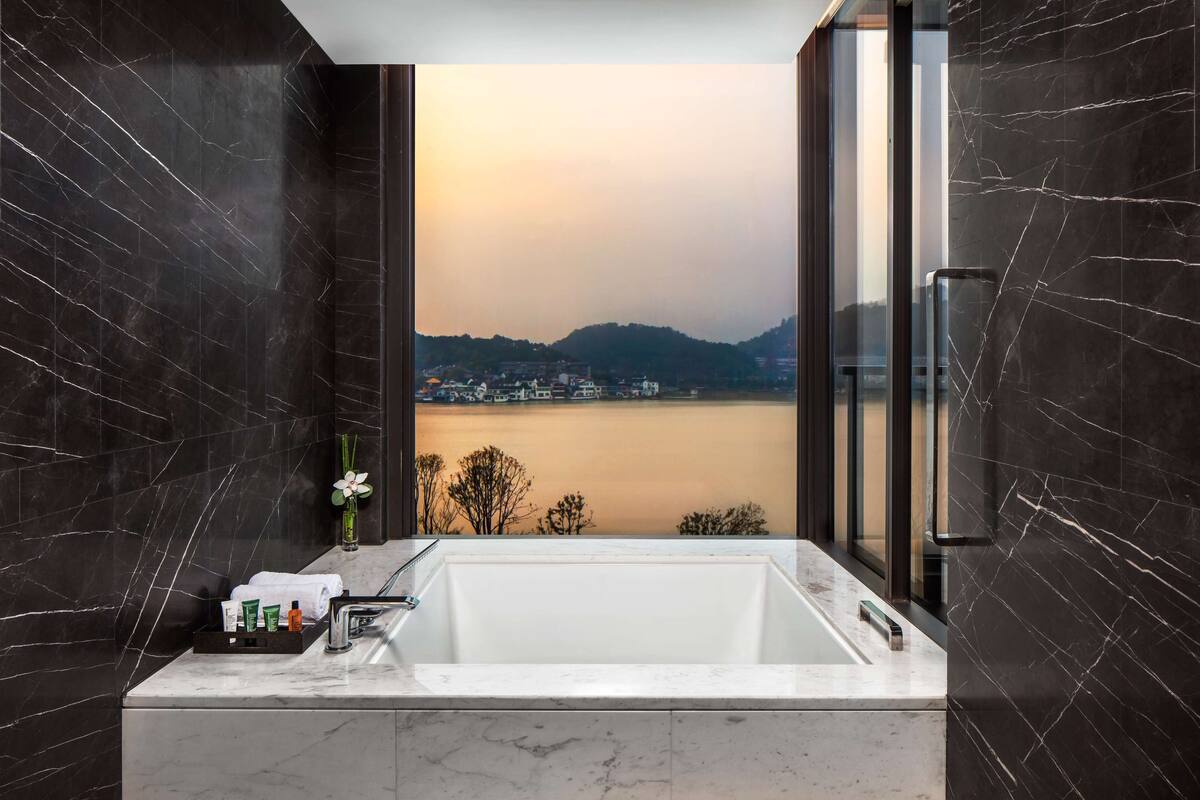Een moderne badkamer met een groot bad, donkere marmeren wanden en uitzicht op water en verre gebouwen.