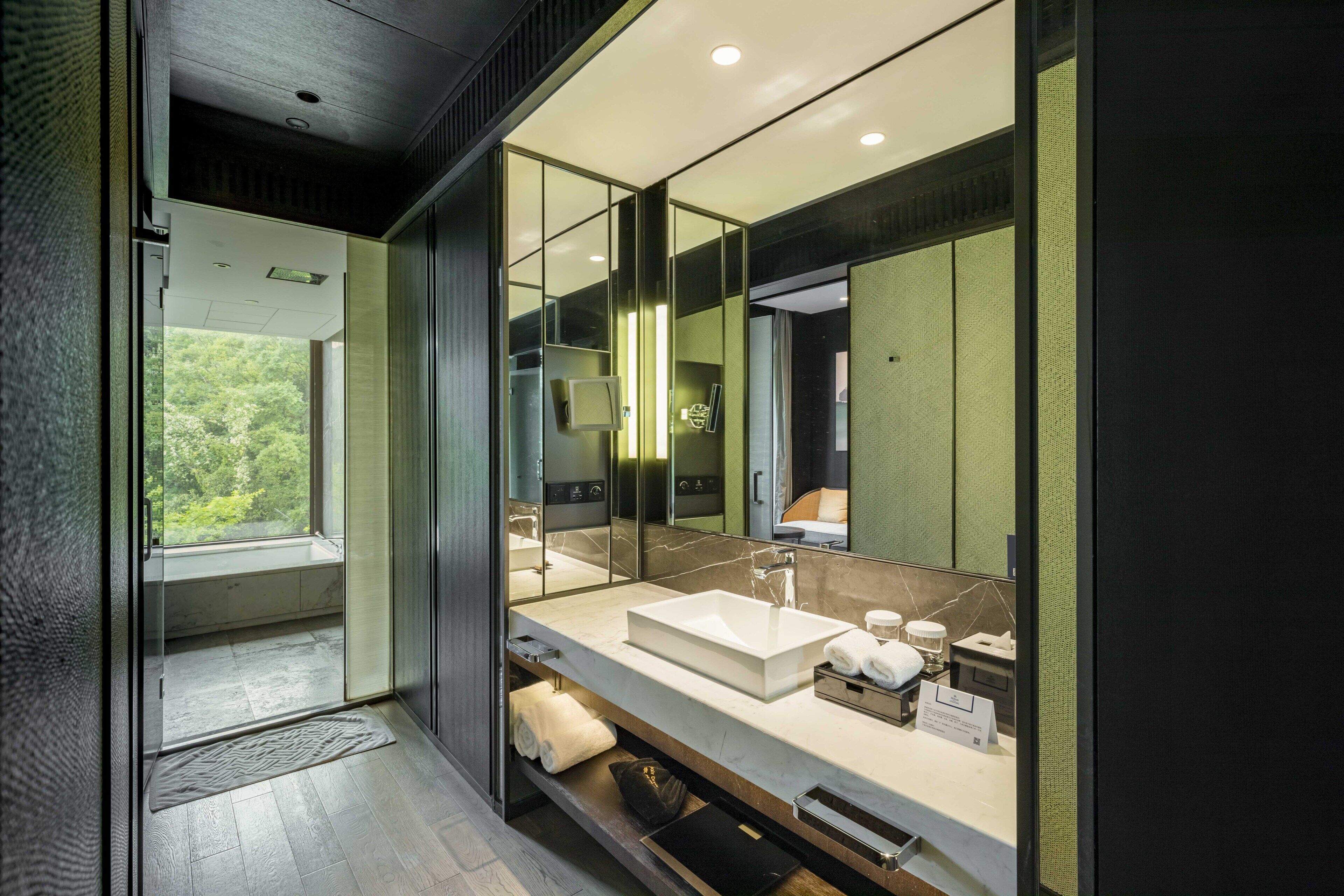 Un baño moderno con un espejo grande, un lavamanos blanco y una ventana que ofrece vistas a la vegetación.
