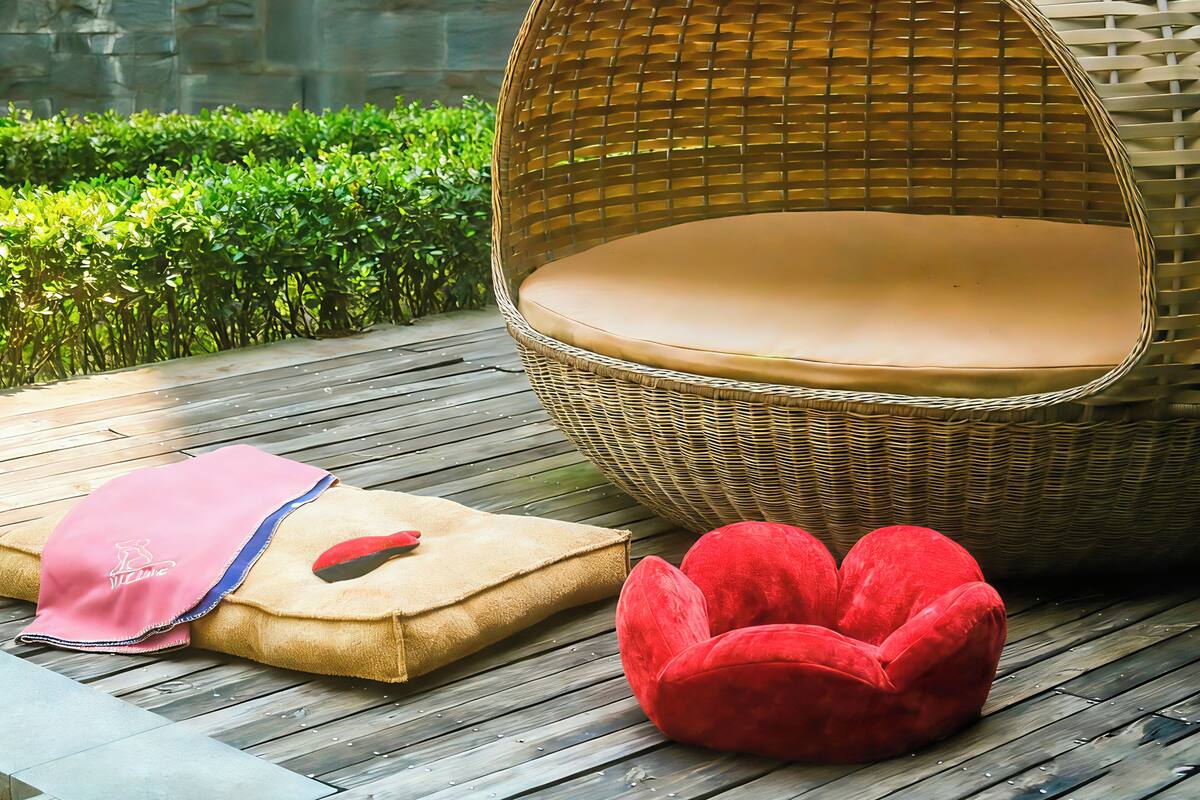 Una silla de mimbre con un cojín, un sillón de judías rojo y un cojín beige con una toalla rosa sobre una terraza de madera.