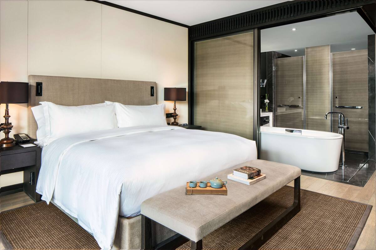 Una habitación de hotel moderna con una cama grande, mesitas de noche, una lámpara de noche, una pequeña mesa con un libro y un calentador de agua, y un baño con bañera y ducha.