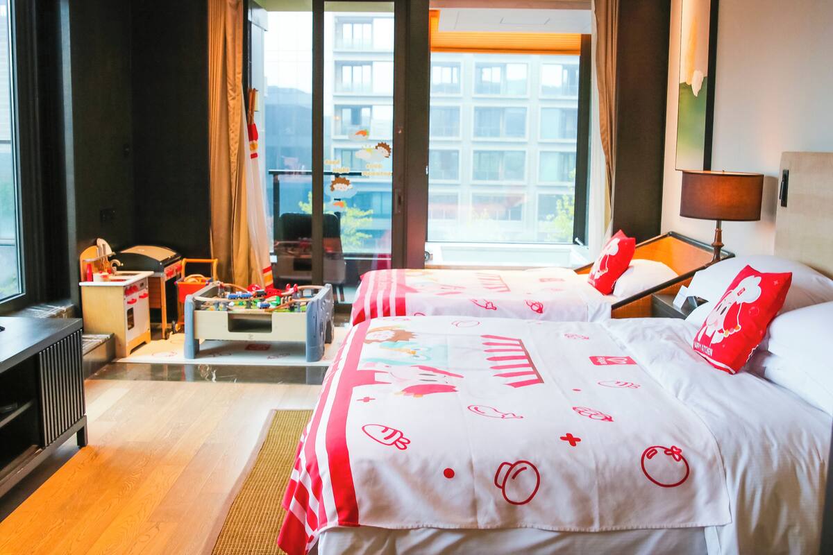 Habitación de hotel con dos camas, un escritorio, una silla, una mesita con juguetes y vistas a edificios a través de un ventanal grande.