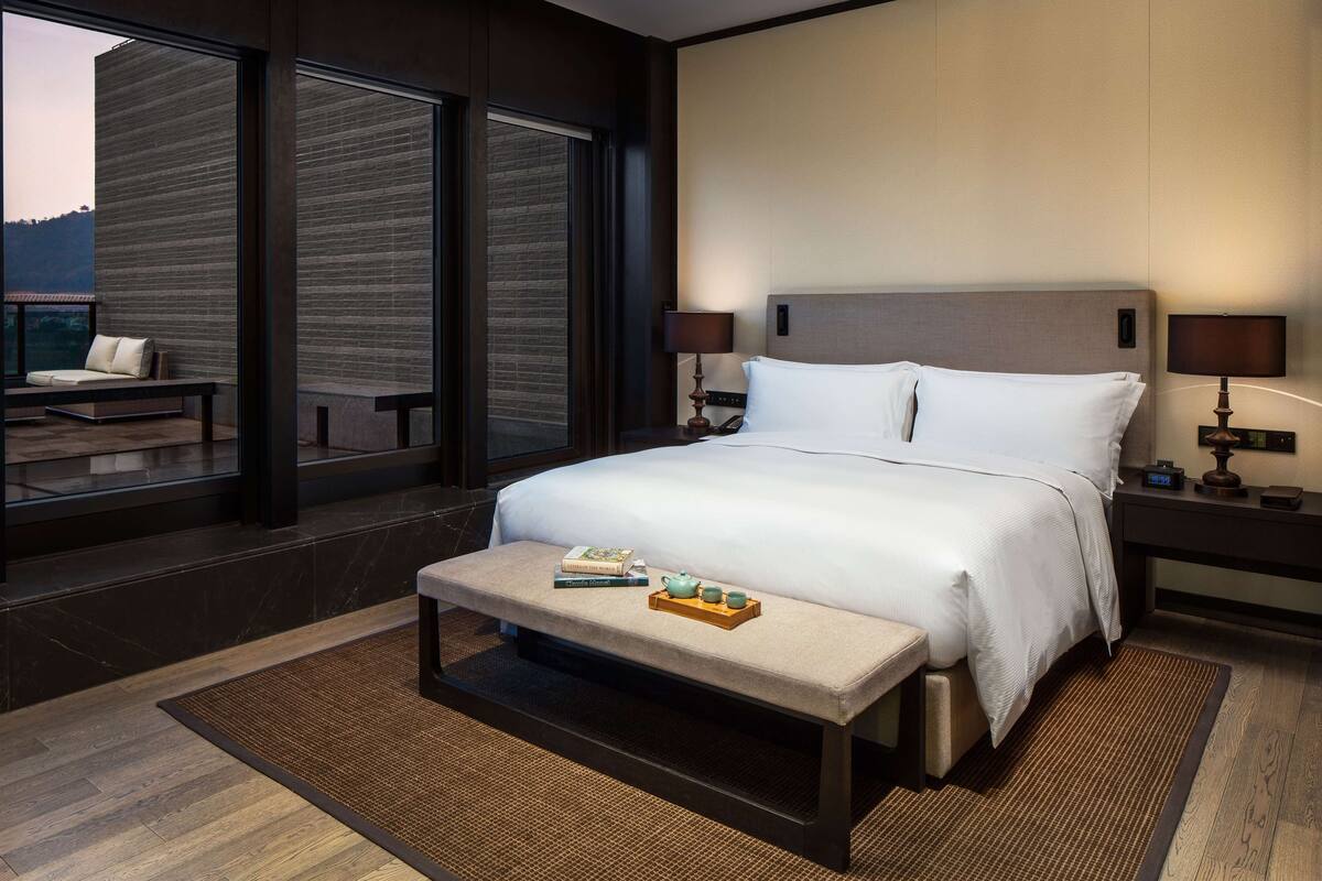 Een moderne hotelkamer met een groot bed, twee nachtkastjes met lampen, een bankje met een dienblad vol boeken en een theepot, en grote ramen met uitzicht op het omliggende landschap.
