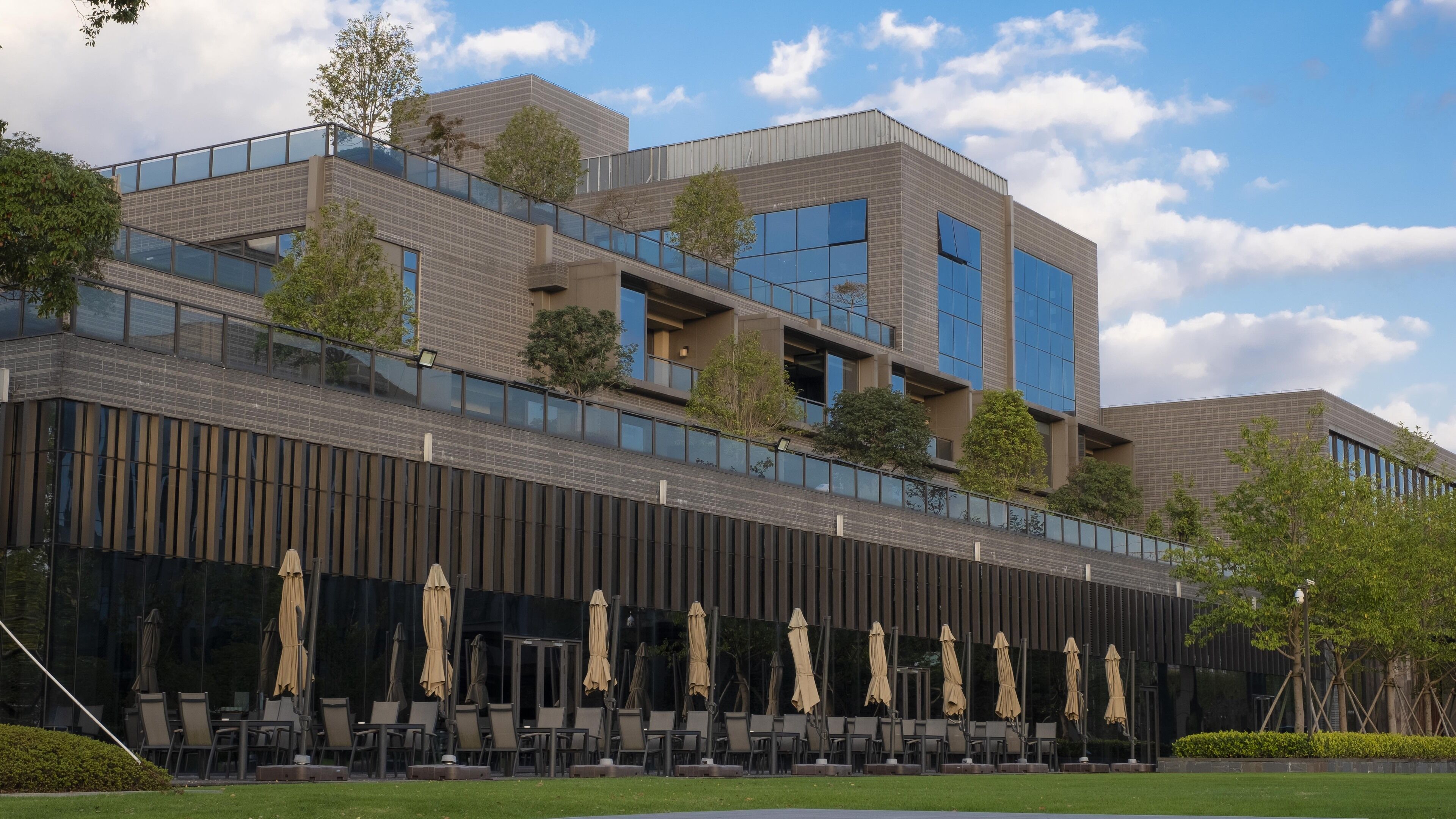 A modern building with a glass facade, outdoor seating area with umbrellas, and a well-maintained lawn.