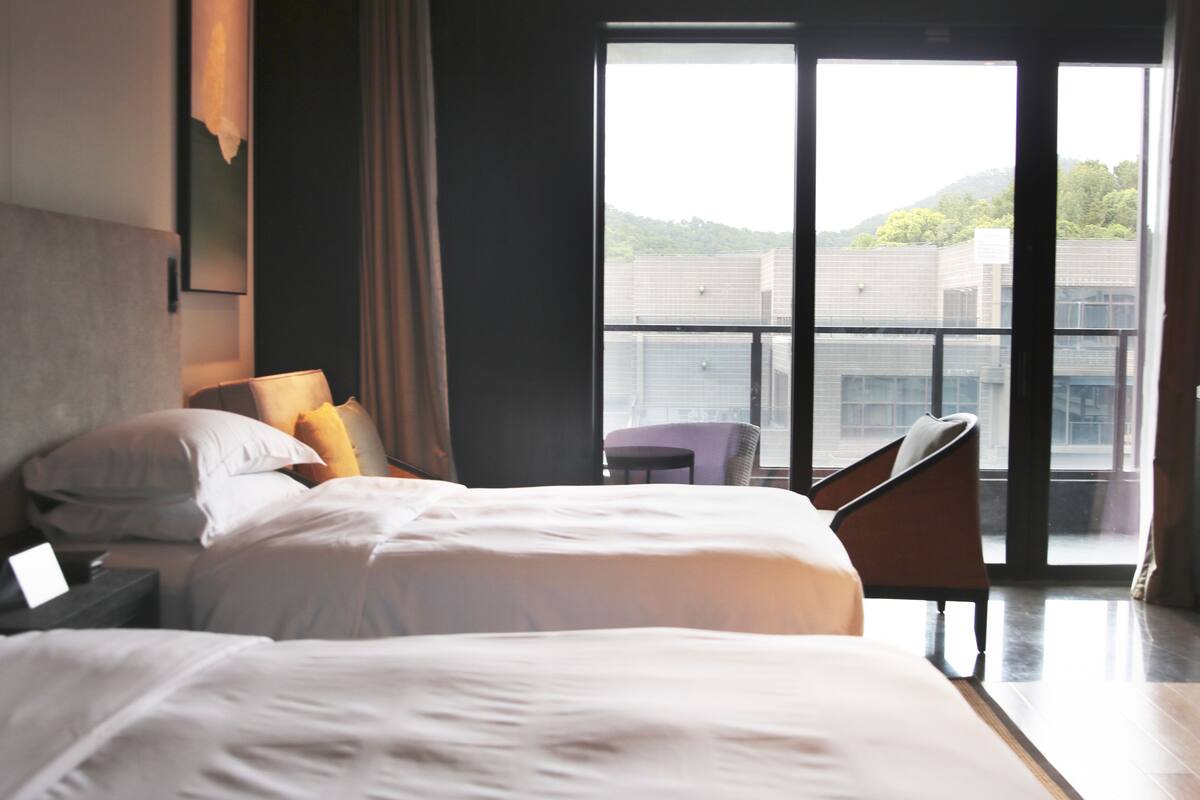 Een hotelkamer met twee bedden, een stoel en een balkon met uitzicht op gebouwen en groen.