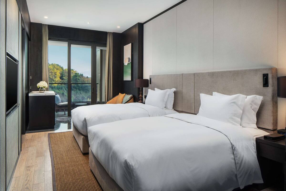 Una habitación de hotel moderna con dos camas, un ventanal con vistas a árboles, una pequeña mesa con un florero y una mesita de noche con una lámpara.