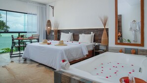 Une chambre d’hôtel dotée d’un grand lit, d’un coin salon séparé avec une table et des chaises, d’une baignoire remplie de pétales de roses et offrant une vue sur la mer grâce à de grandes fenêtres.