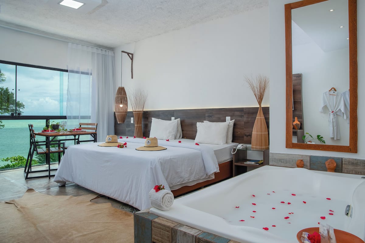 Habitación de hotel con una cama grande, un área de estar separada con una mesa y sillas, una bañera con pétalos de rosa y vistas al mar a través de amplias ventanas.