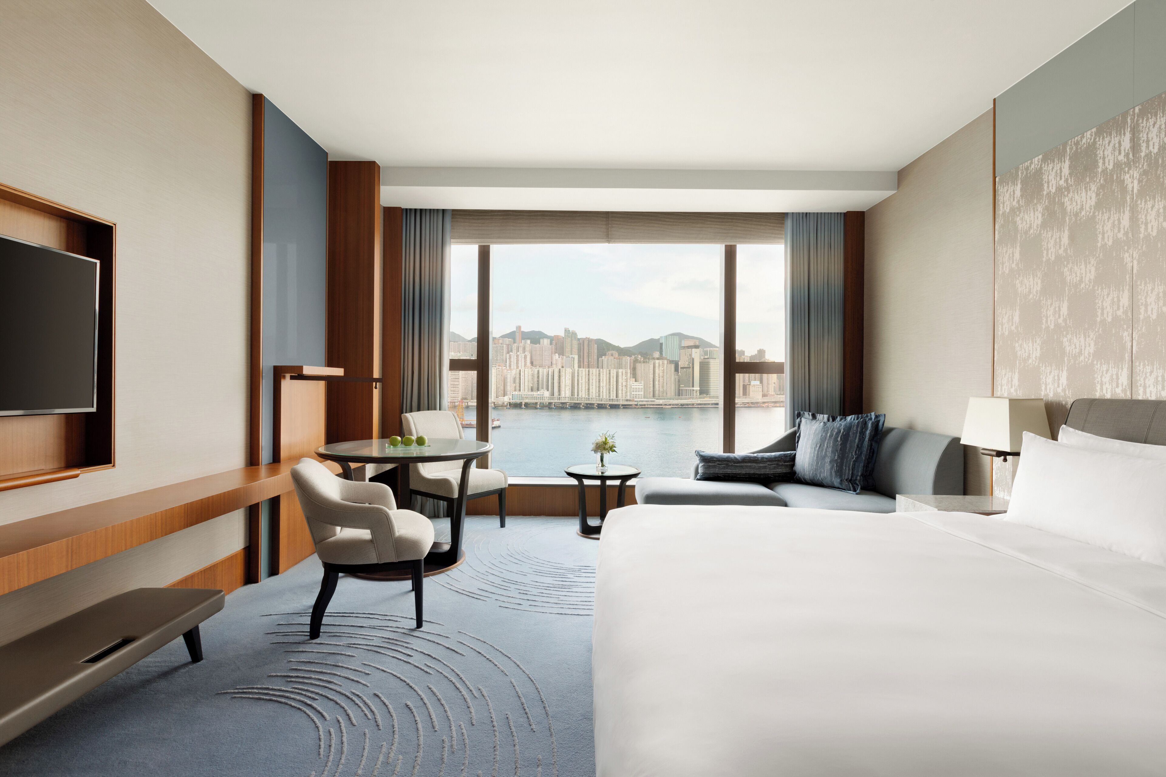 A hotel room with a large bed, a desk, a chair, a small table, and a TV. There is a view of a cityscape through large windows.