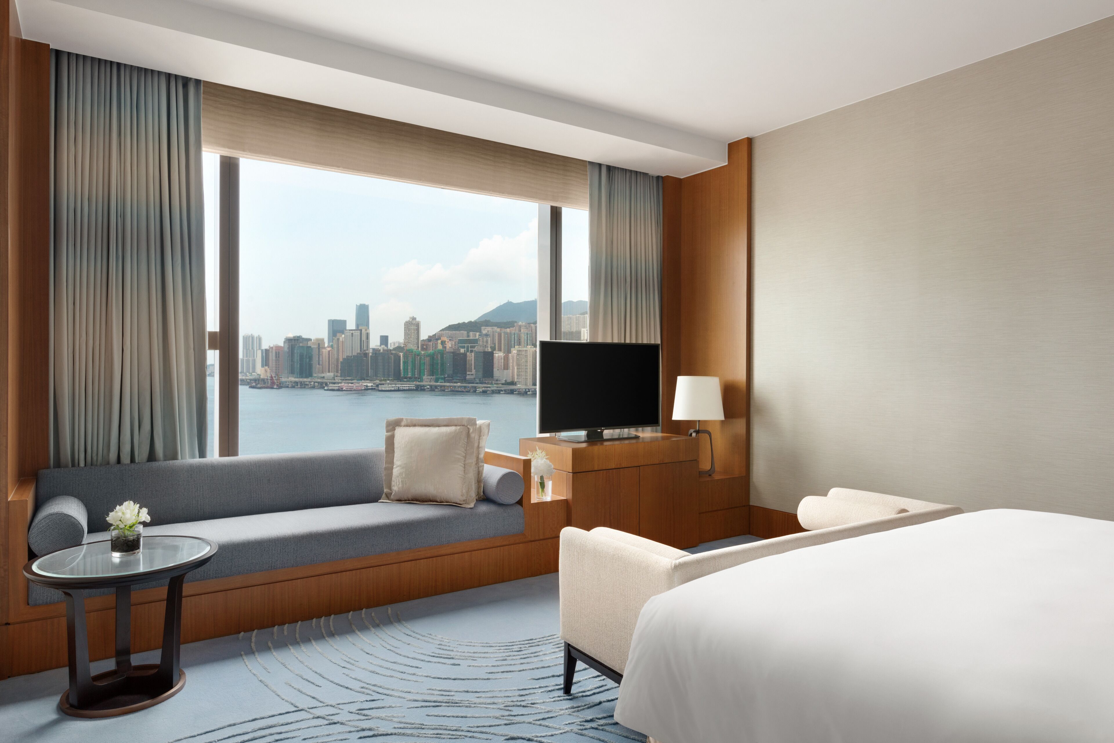 A hotel room with a large window, a sofa, a bed, a TV, and a desk with a lamp.