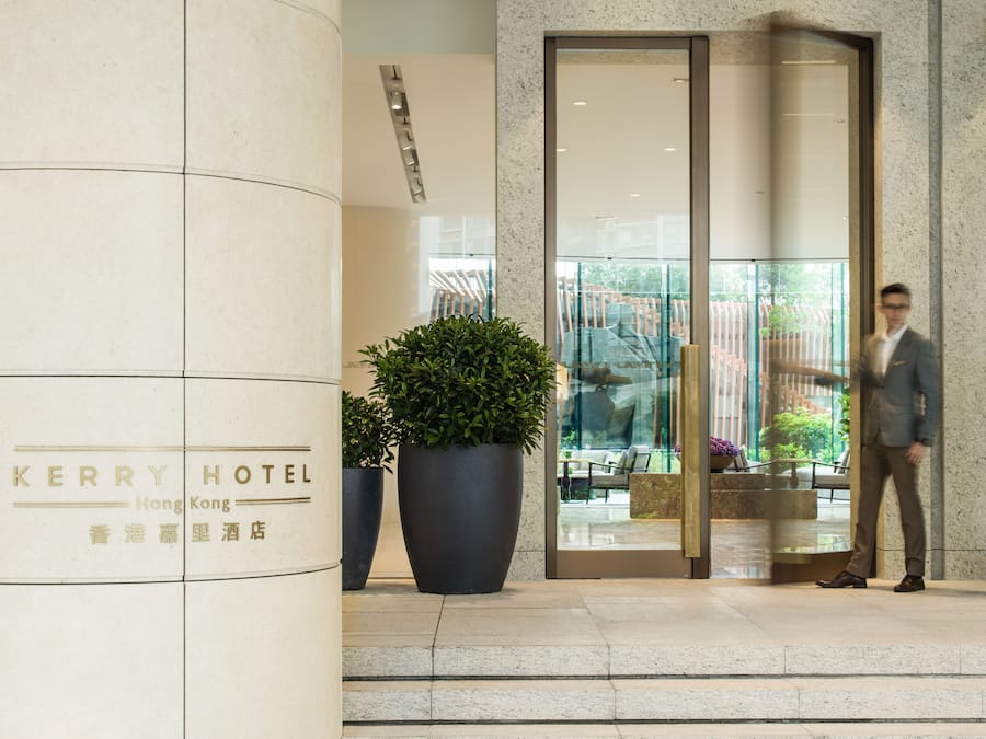 A hotel entrance with a large potted plant, a glass door, and a man in a suit standing by the door.