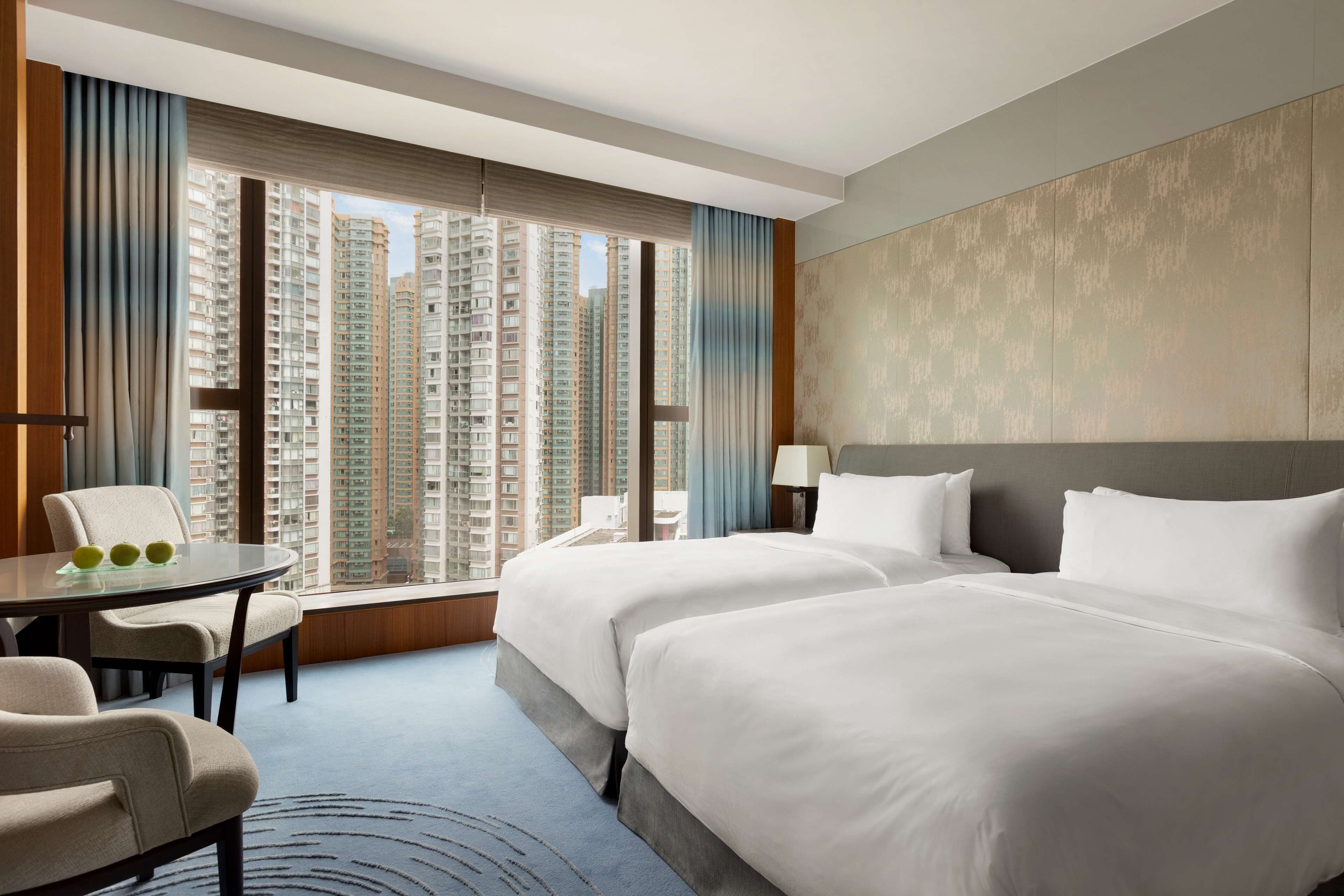 A hotel room with two beds, a dining table, and a view of high-rise buildings.