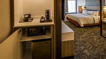 A hotel room with a compact kitchenette, a bed with a headboard, and a view of the outside.