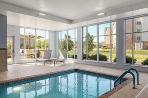 Una piscina con agua azul cristalina, rodeada de amplias ventanas que ofrecen vistas a un jardín ajardinado y edificios.