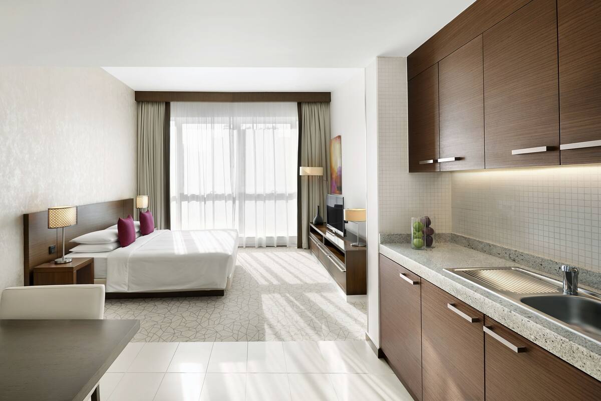 A modern hotel room with a bed, bedside tables, a desk, a TV, and a kitchen area with a sink and cabinets.