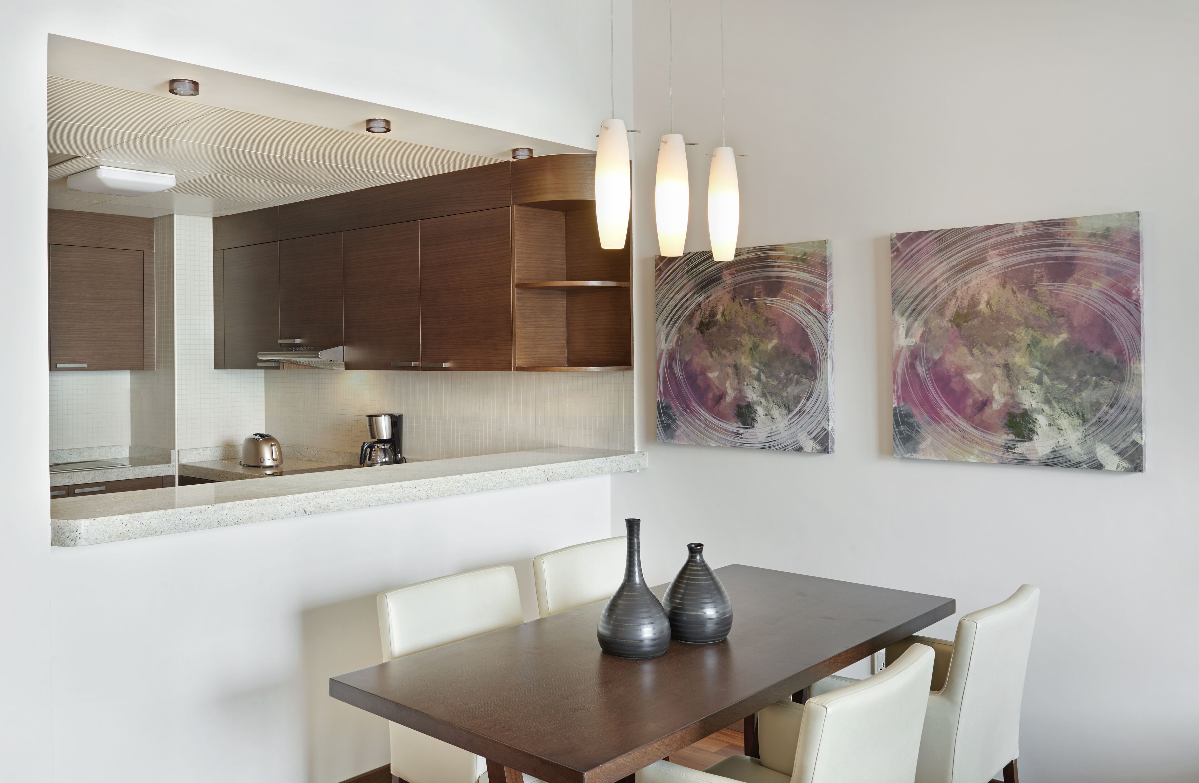 A modern kitchen with wooden cabinets, a countertop, and a dining area with a table and chairs. There are two abstract paintings on the wall.