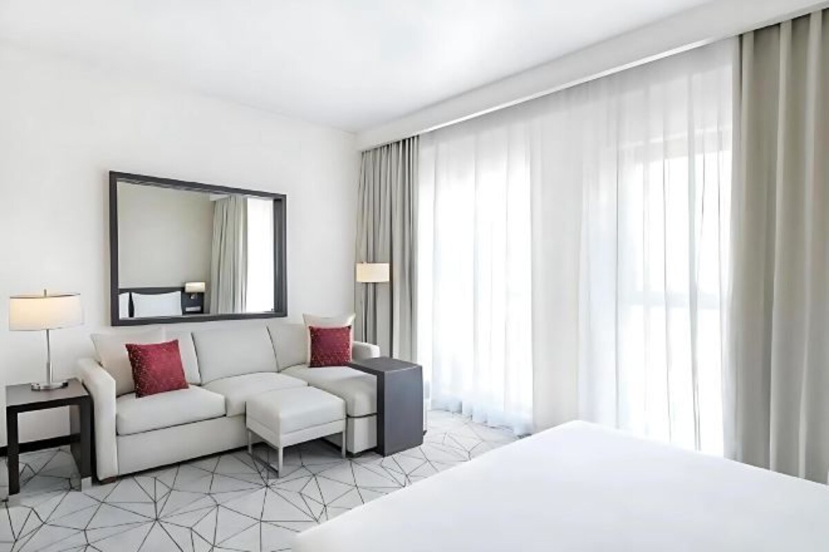 Une chambre d’hôtel avec un lit, un canapé, un bureau, une chaise, un miroir et une fenêtre avec des rideaux.