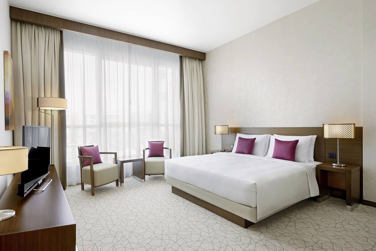 A hotel room with a large bed, two bedside tables, a desk with a TV, a chair, and a large window with curtains.