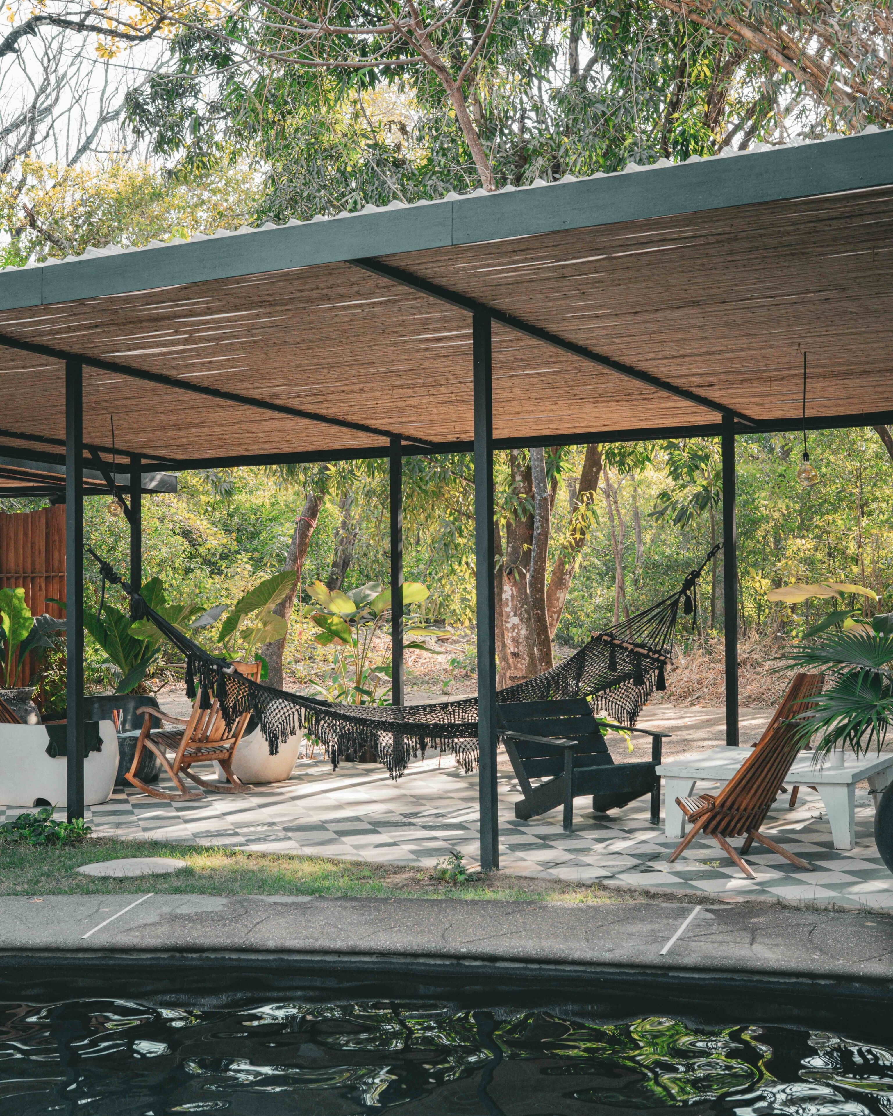 A covered outdoor seating area with hammocks and chairs, surrounded by trees and a water feature.