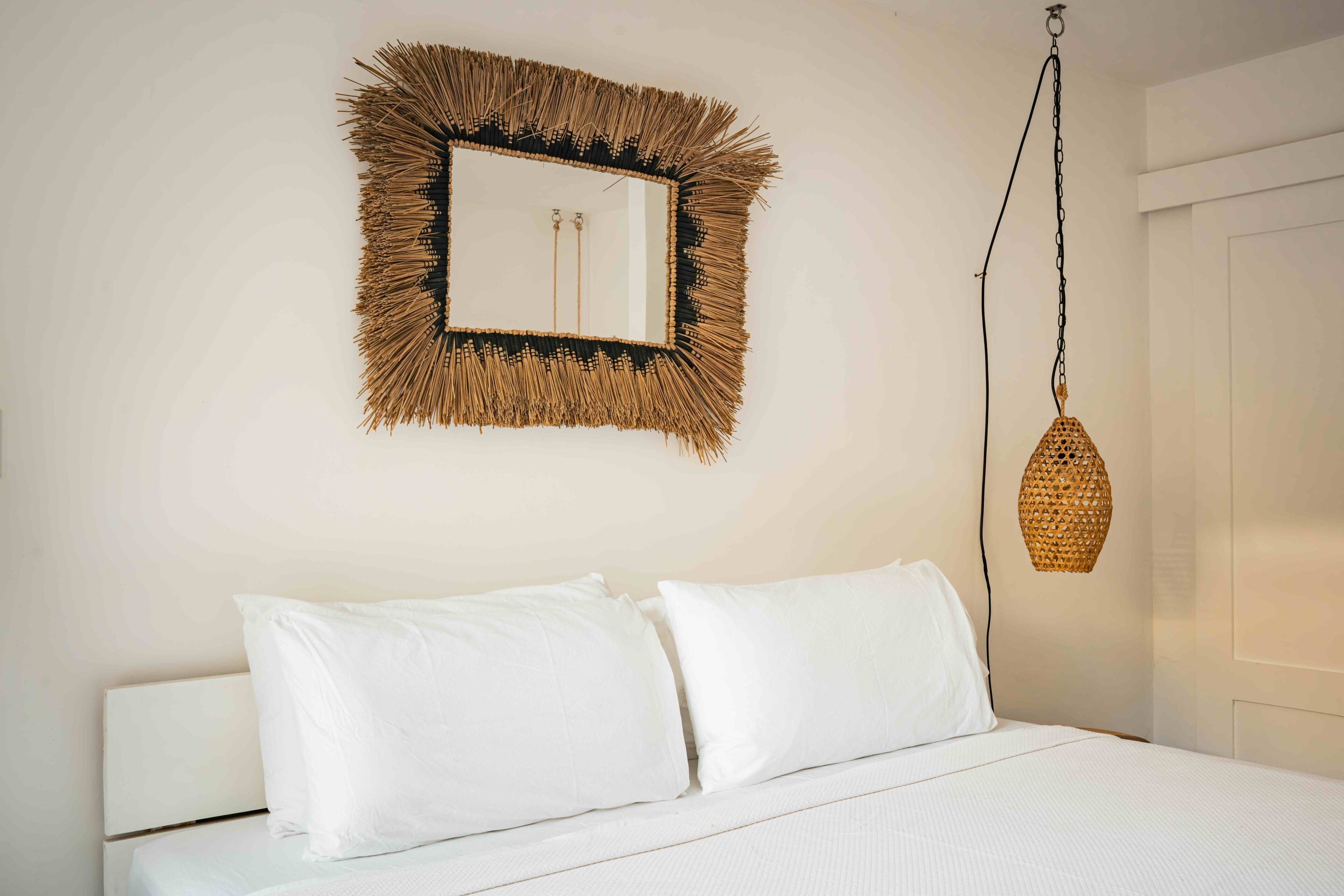 A bed with white bedding, a wall-mounted mirror with a natural frame, and a hanging wicker light fixture.