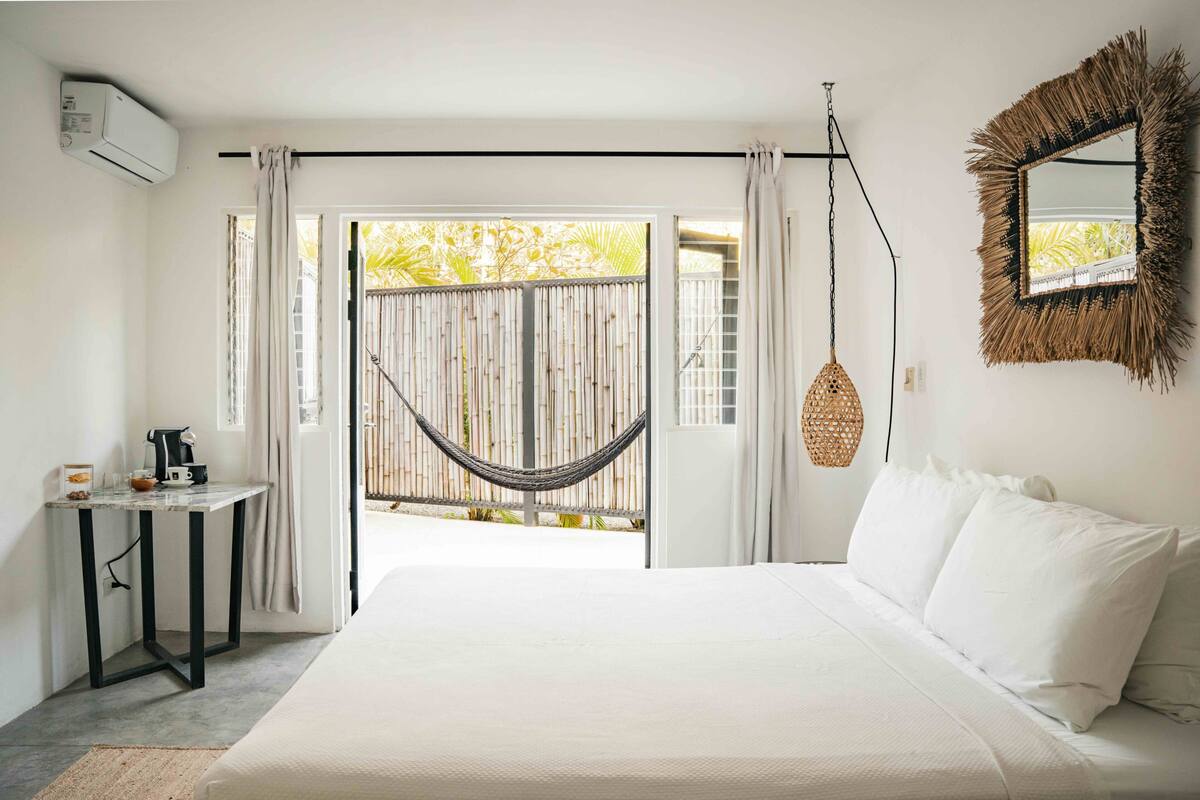 Une chambre avec un lit, une petite table, une lampe suspendue et une vue sur l’extérieur, visible à travers une porte.