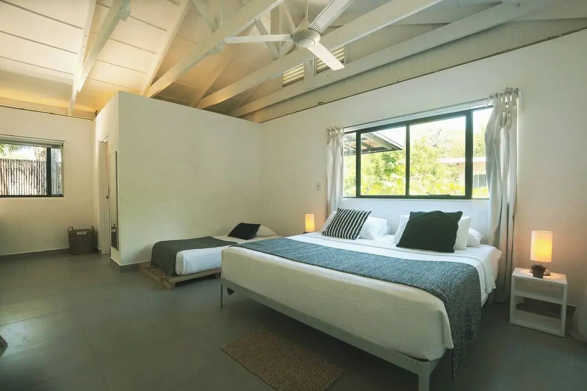Une chambre avec un grand lit, un lit plus petit, une fenêtre avec des rideaux, une lampe de chevet et un ventilateur de plafond.