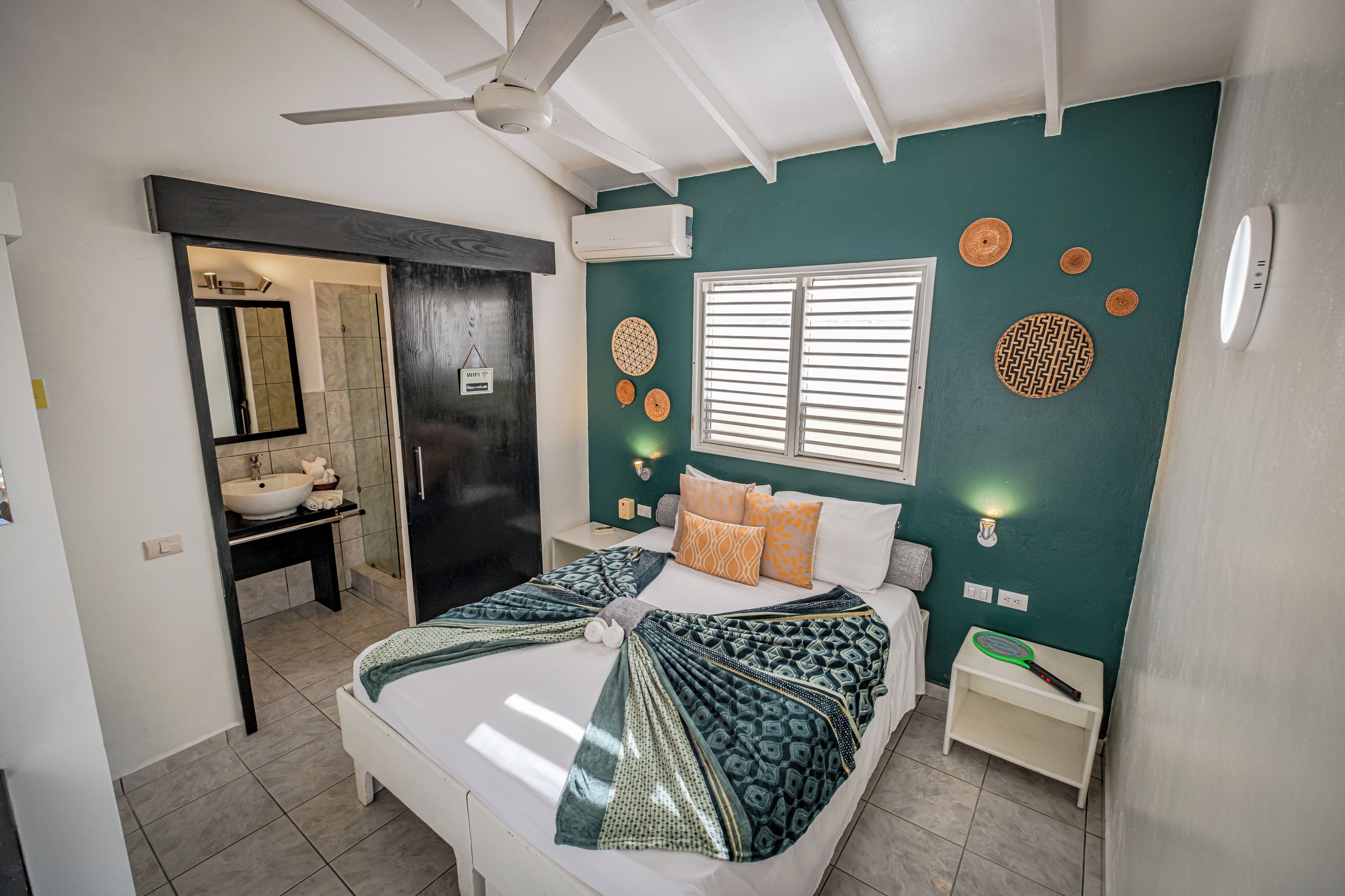 Une chambre à coucher comprenant un lit, une table de chevet, une lampe fixée au mur, une fenêtre avec des stores et un ventilateur de plafond.