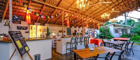 A restaurant with a rustic thatched roof, wooden tables and chairs, a bar area with a chalkboard menu, and a chalkboard sign with services listed.