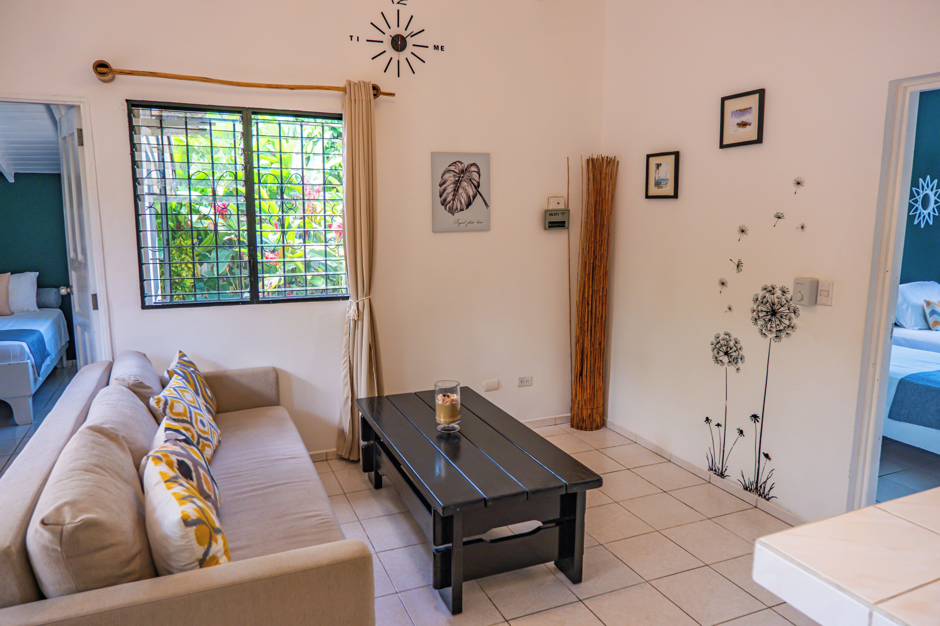 Una sala de estar con un sofá, una mesa de centro, una ventana con cortinas, un reloj de pared y una puerta que da a otra habitación.