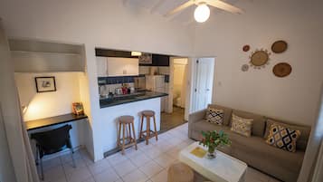 A modern kitchen with a central island, a dining area with stools, a living room with a sofa and cushions, and a ceiling fan.