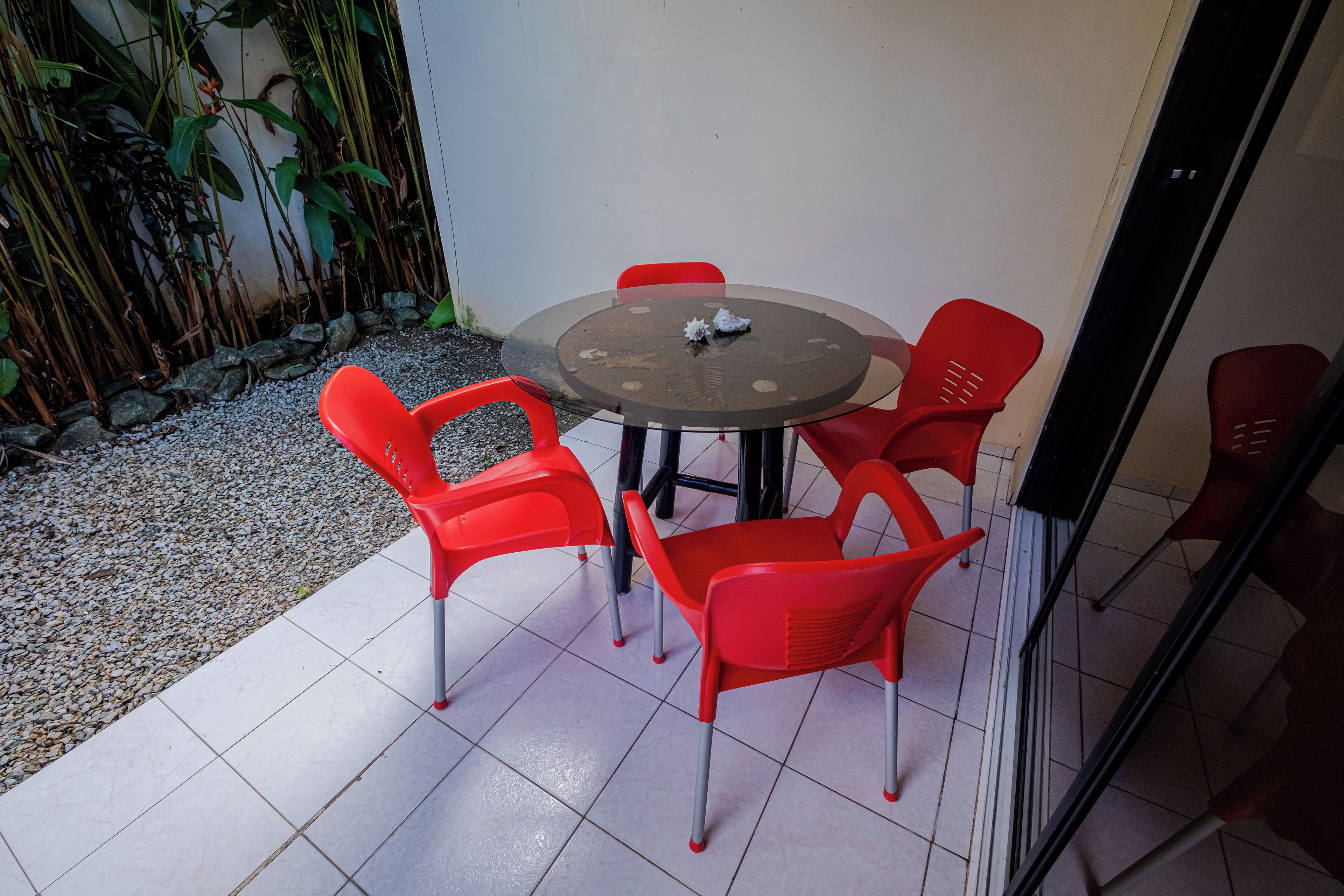 A round glass table with four red plastic chairs, surrounded by a gravel area and a bamboo fence.