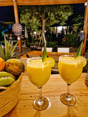 Två drinkar med limeskivor som garnering står på ett träbord, en korg med frukt och en darttavla syns i bakgrunden.