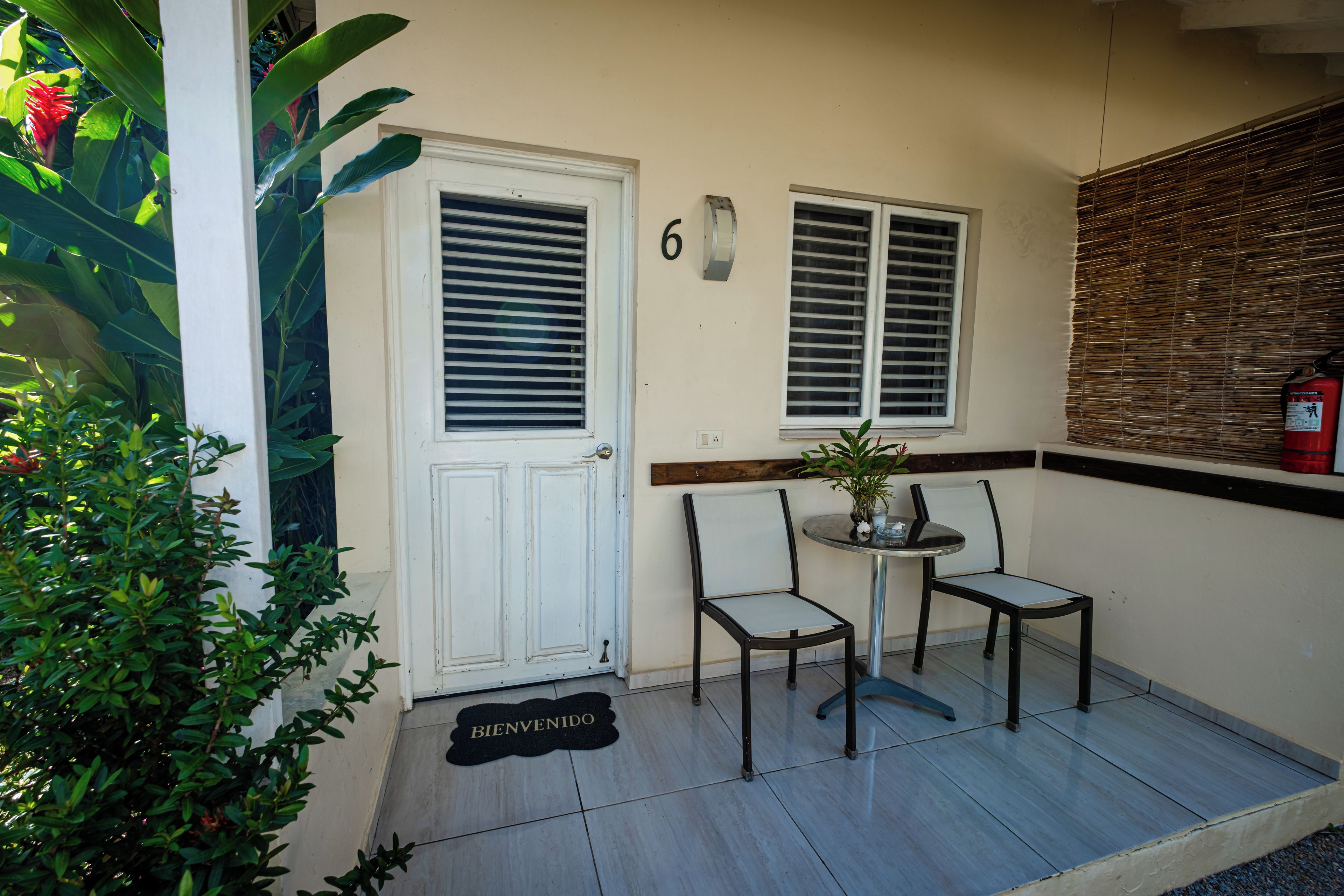Une entrée propre et bien entretenue, avec une porte, deux fenêtres et une petite table avec des chaises. Un paillasson est posé sur le sol, et un extincteur est fixé au mur.
