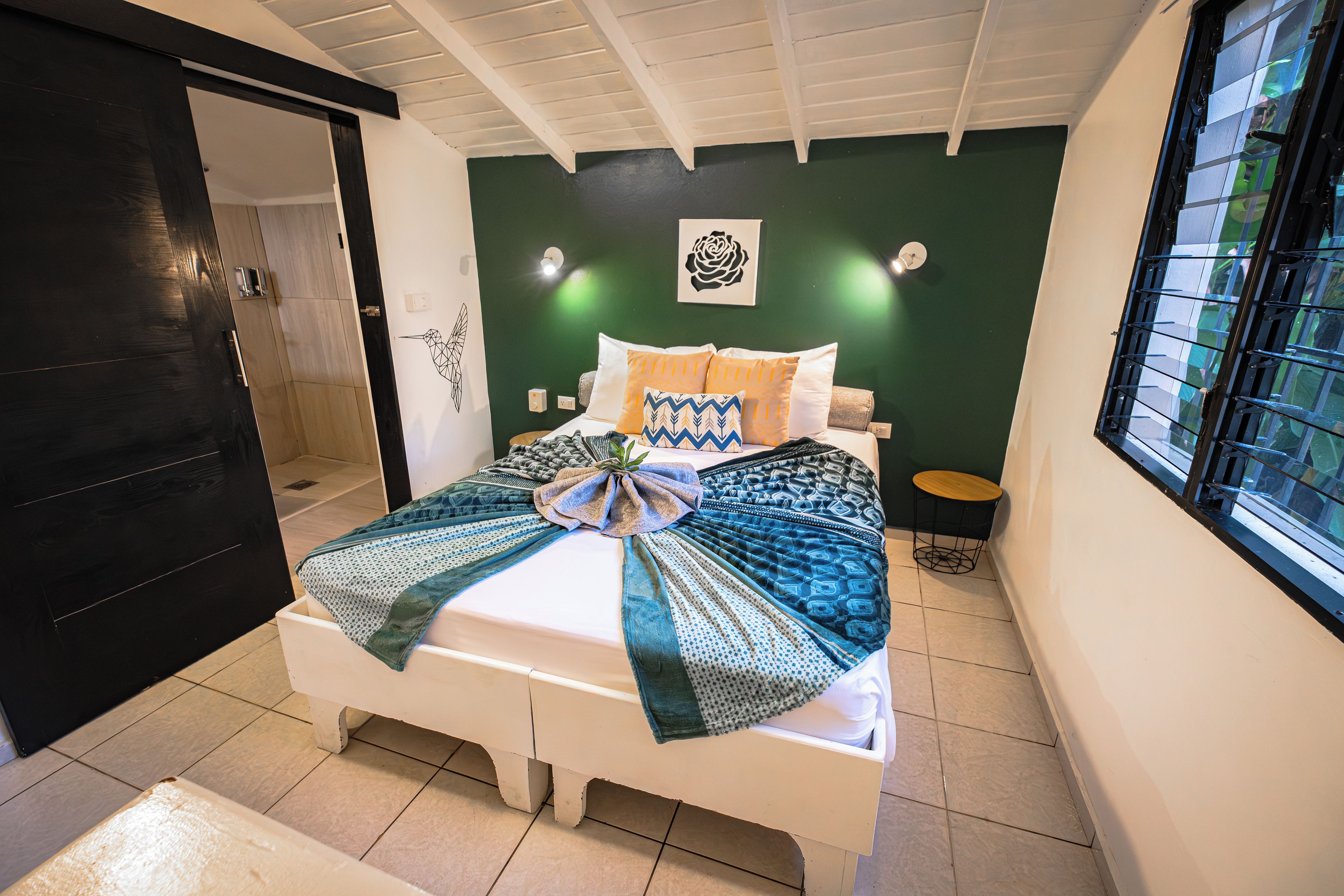 A bedroom with a bed, a small bedside table, a wall-mounted light, and a window with a metal grill.