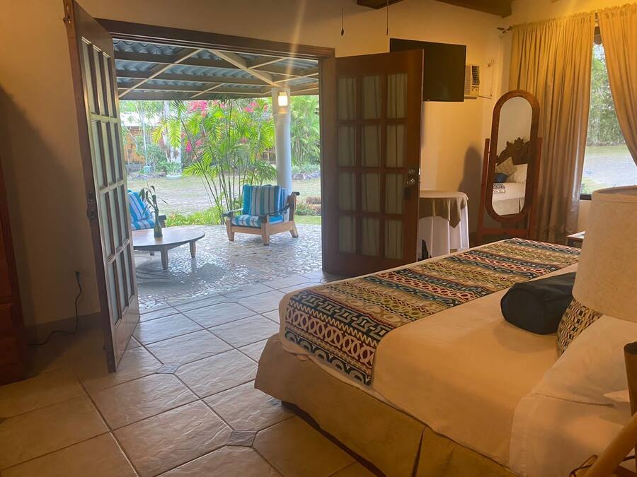 A hotel room with a bed, a small table, a chair, and a view of a garden through an open door.