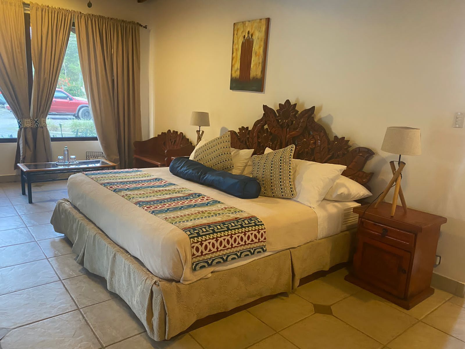 A hotel room with a large bed, a wooden headboard, two bedside tables with lamps, a window with curtains, and a painting on the wall.