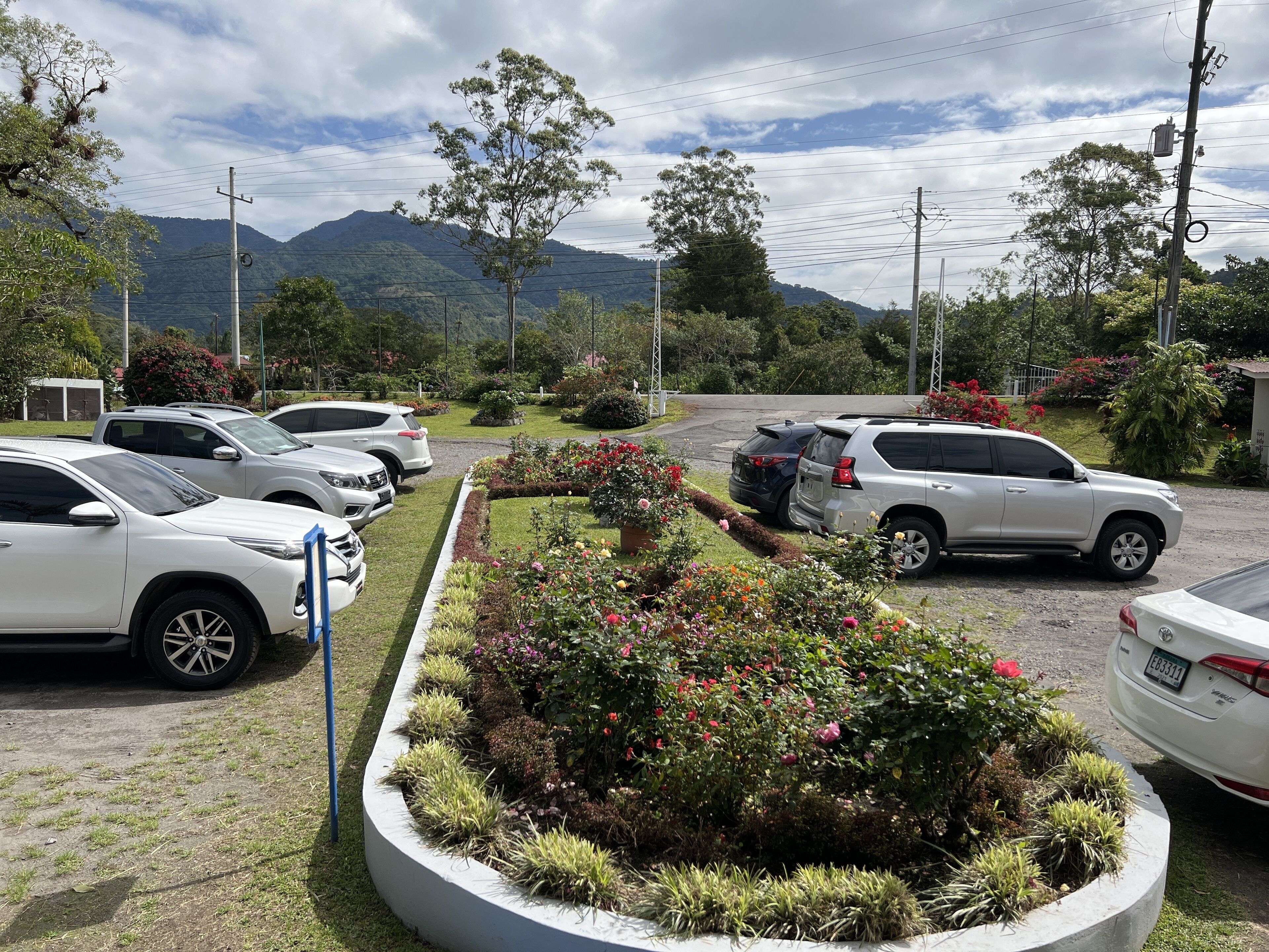 Un estacionamiento con varios coches, un jardín ajardinado con diversas plantas y flores, y un paisaje montañoso al fondo.