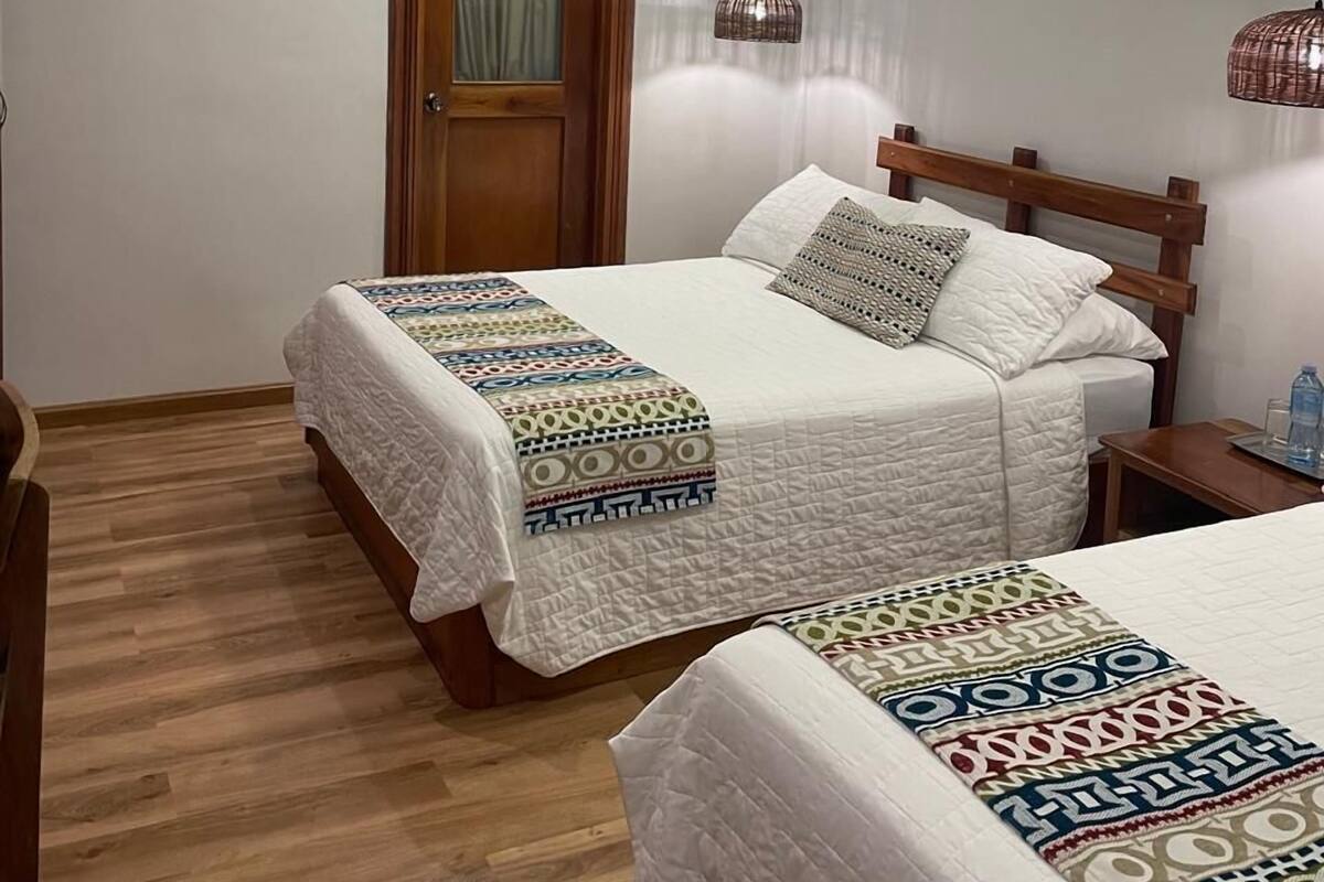 A hotel room with two beds, wooden headboards, a wooden door, a ceiling-mounted air conditioner, and a hanging pendant light.