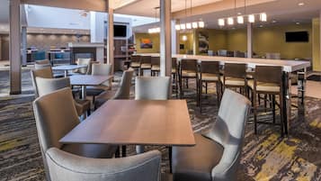 Um lobby de hotel moderno com uma área de refeições central, diversas opções de assento e um bar visível.