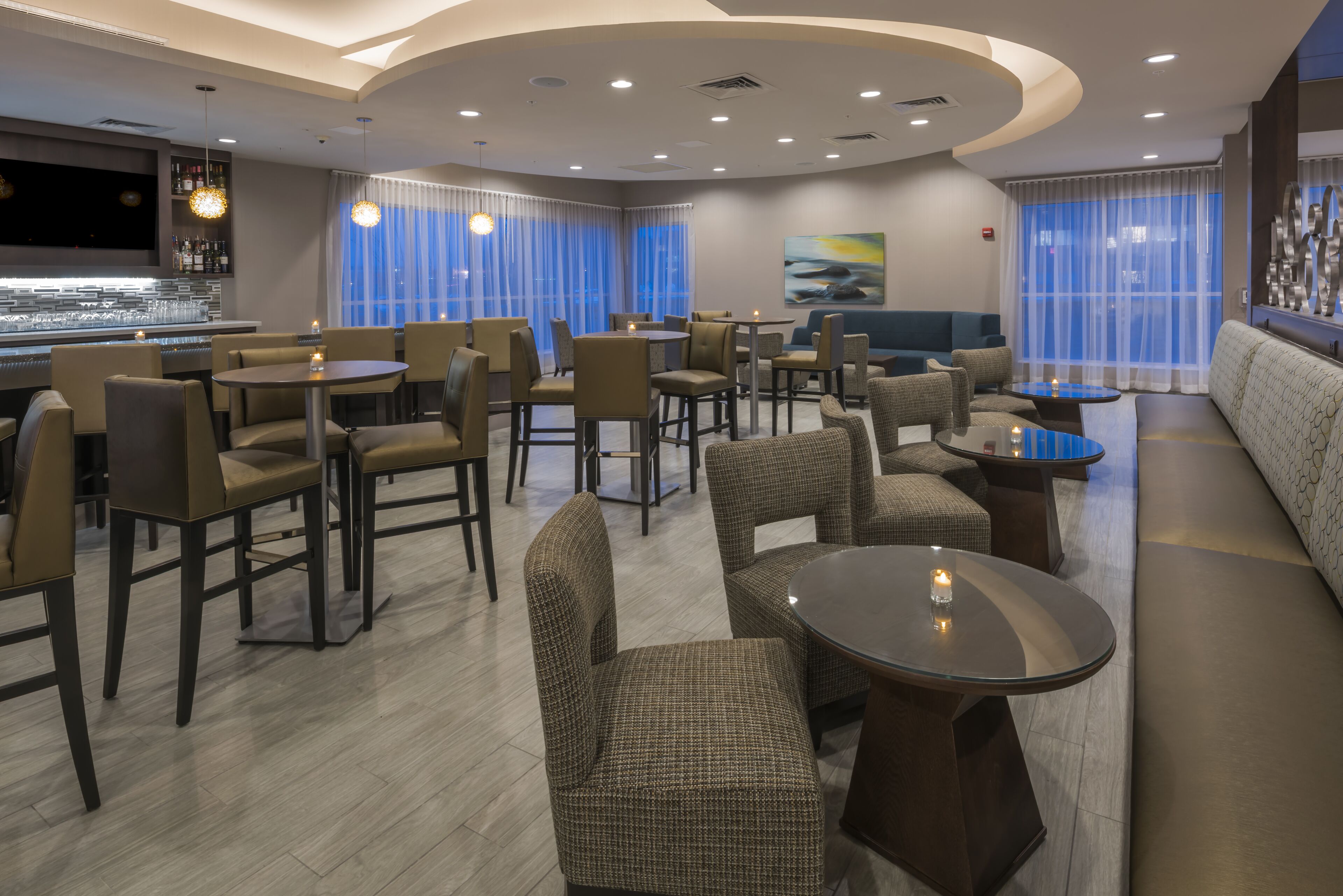 Um saguão de hotel moderno, com área de bar, espaços de estar e uma grande janela com cortinas.