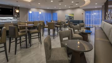 Um saguão de hotel moderno, com área de bar, espaços de estar e uma grande janela com cortinas.