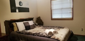 Un dormitorio con cama, almohadas, una manta, un oso de peluche, una ventana y un cuadro en la pared.