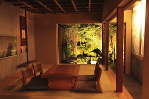 Um quarto japonês tradicional com tatames, uma mesa de madeira baixa e cadeiras. Uma porta deslizante dá acesso a um jardim com lago e árvores.