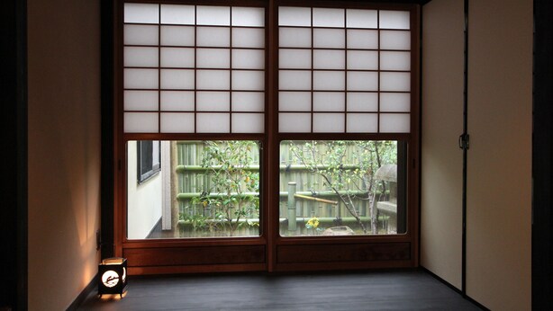 Una habitación tradicional japonesa con puertas correderas, una pequeña mesa con un reloj y vistas a una valla de bambú y follaje exterior.