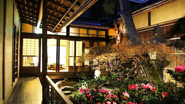 Un interior tradicional japonés con suelo de tatami, escalera de madera y vistas a un jardín con flores rosadas.