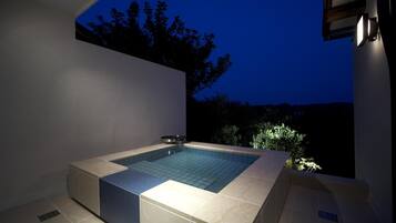 A modern outdoor hot tub area with a tiled floor, surrounded by walls and a view of trees and hills at dusk.