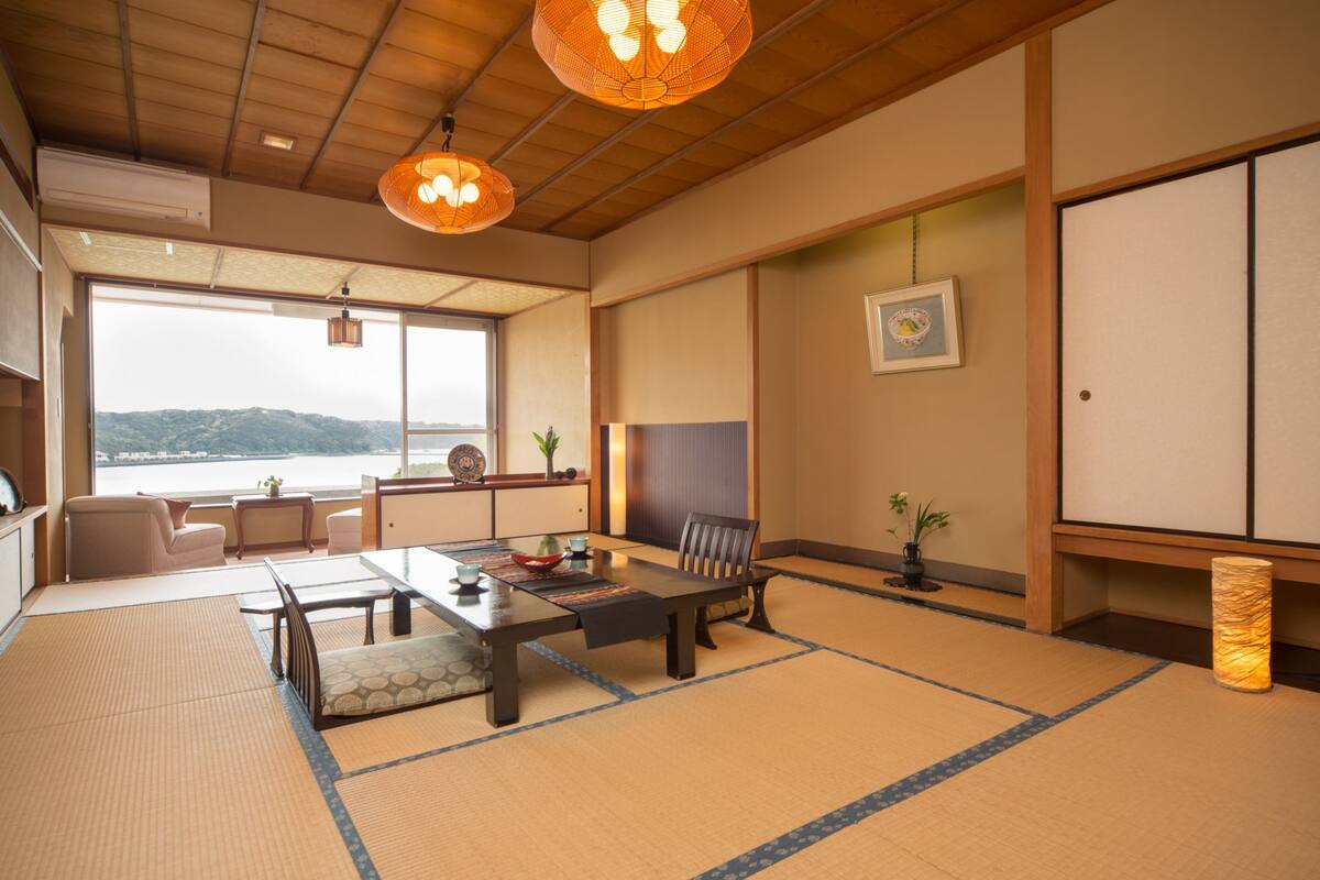 A traditional Japanese room with tatami flooring, a low table with a tea set, a cushioned chair, and a view of a body of water.