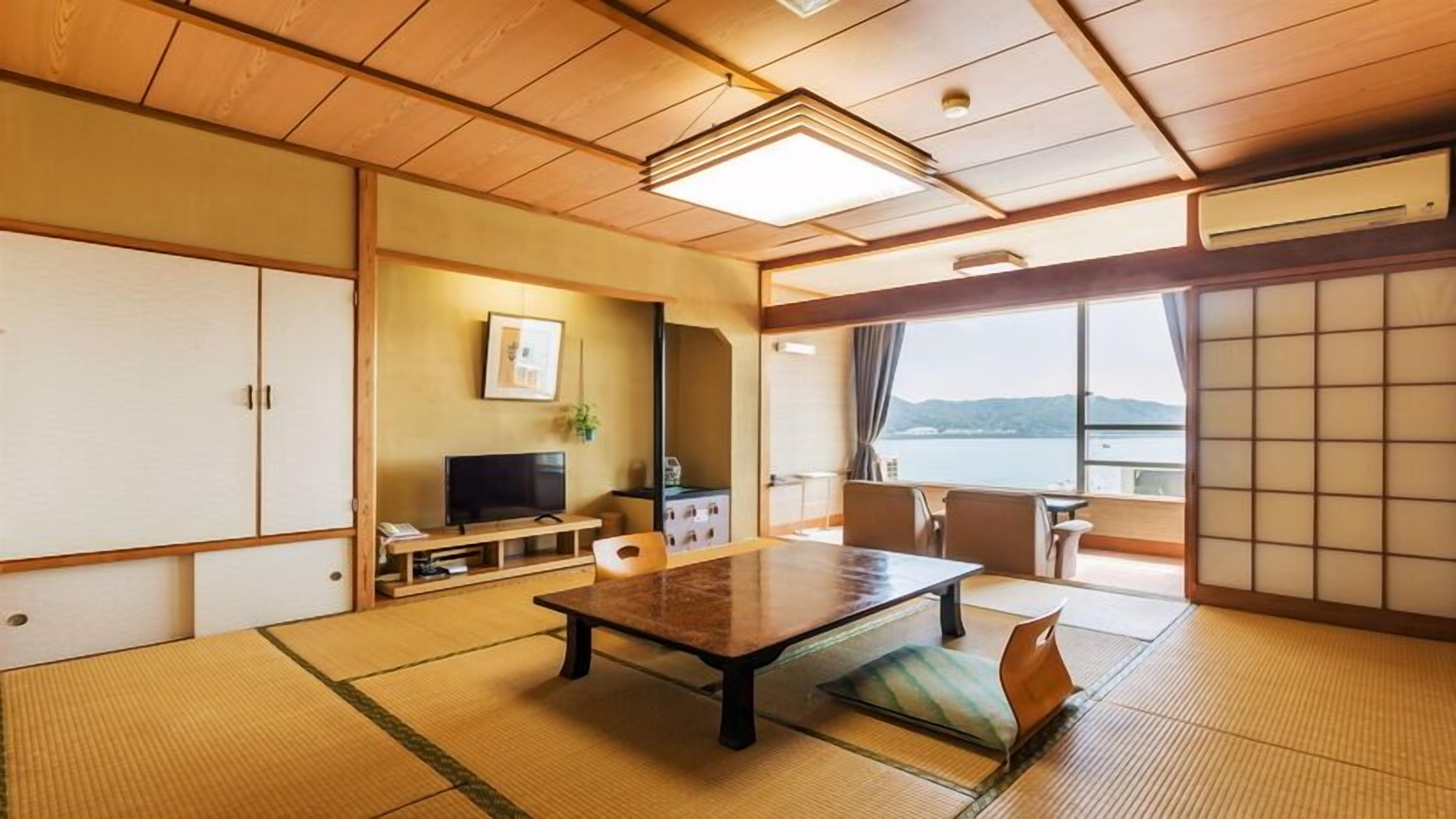 A traditional Japanese room with tatami flooring, a low table, a television, and a view of the water.