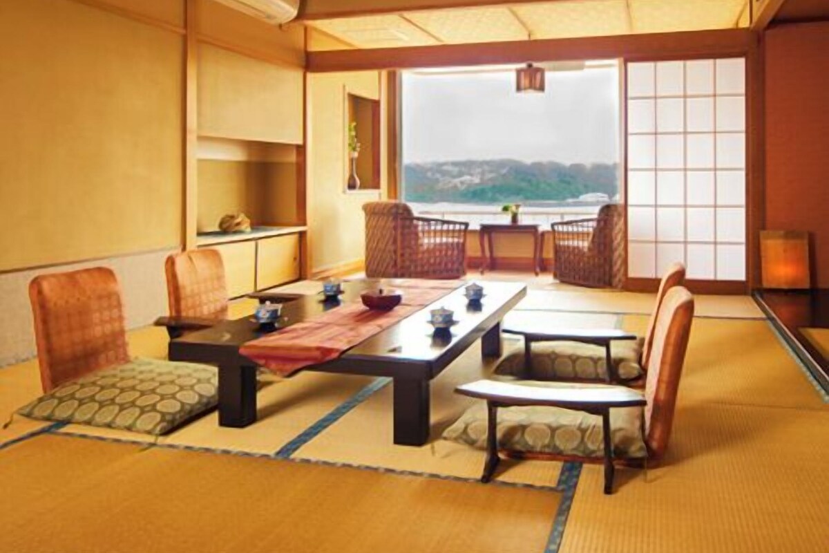 A traditional Japanese-style room with tatami flooring, a low table with cushions, and a view of a body of water.